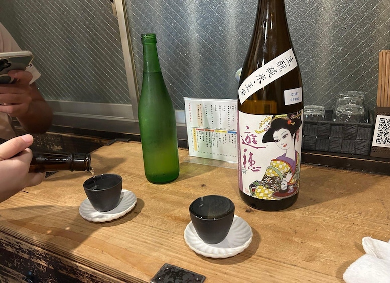 En sakeflaske med traditionel japansk etiket, to små sorte kopper og en grøn flaske på et træbord. 4