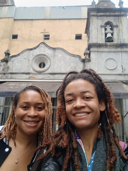 Two people in front of a building with a bell tower and a weathered facade. 1
