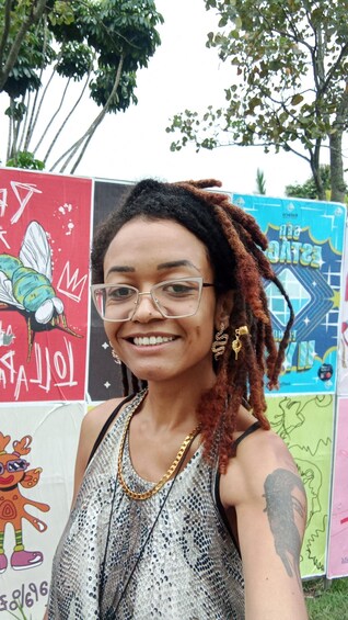 A person with dreadlocks, wearing a sequined top, standing in front of colorful graffiti art. 2