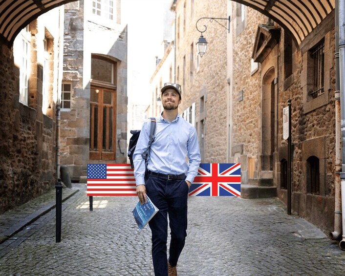 A man walking down a cobblestone street with stone buildings, an American flag, and a British flag in the background. 1