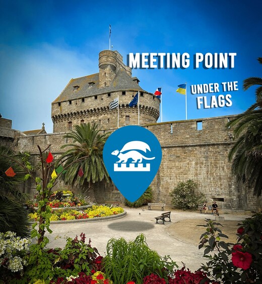 A historic stone building with flags, a blue location marker, and a garden with flowers. 2