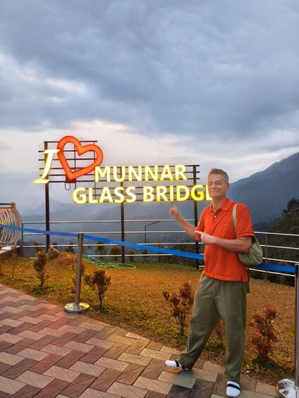 A person standing in front of a sign that reads 'I ♥ MUNNAR GLASS BRIDGE' with a mountainous backdrop. 5