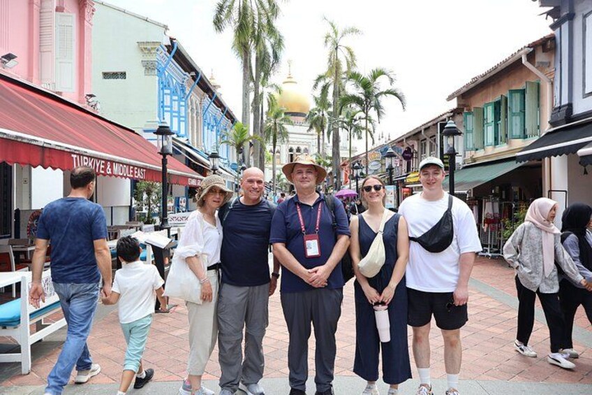 A group of people standing on a pedestrian street with buildings and palm trees in the background. 1