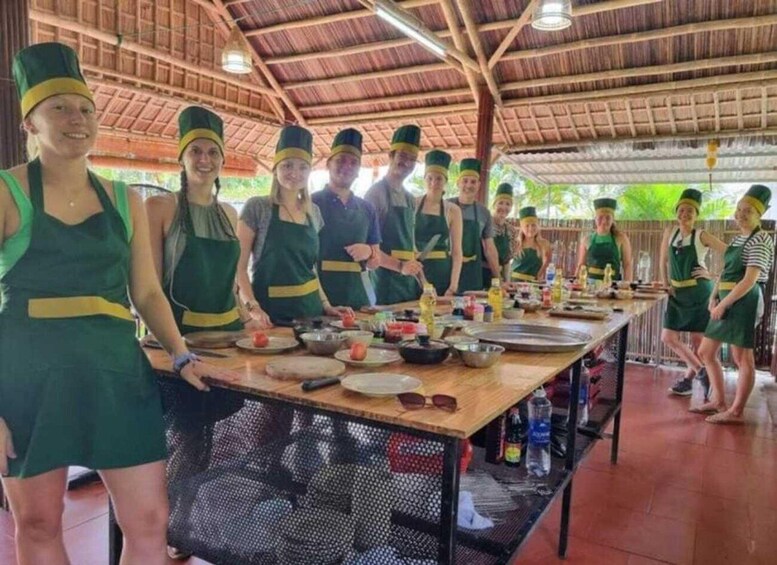 A group of people in green aprons and hats preparing food at a long table in a covered outdoor area. 3