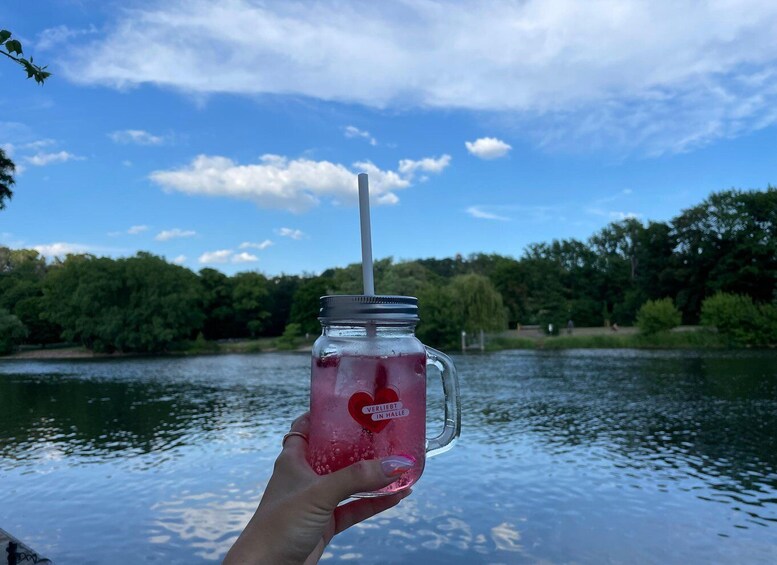 A hand holding a mason jar with a straw, filled with a pink beverage, against a backdrop of a lake and trees under a blue sky. 3