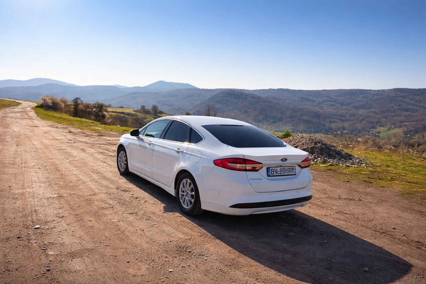 A white sedan parked on a dirt road with a mountainous landscape in the background. 4