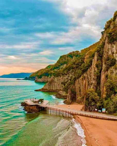 A coastal landscape with a sandy beach, a wooden pier, and steep cliffs covered in greenery. 5