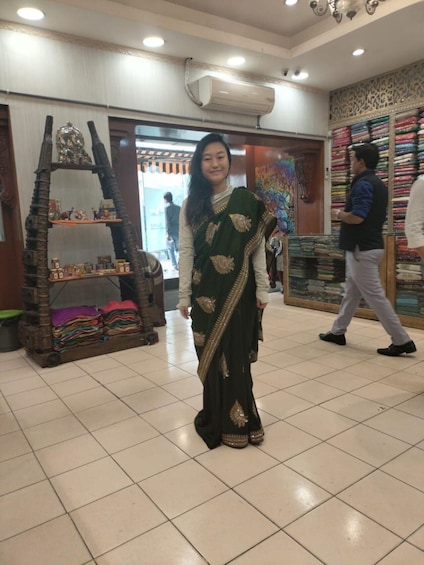 A person in a green saree standing in a shop with shelves of colorful fabrics and a decorative shelf with items. 5