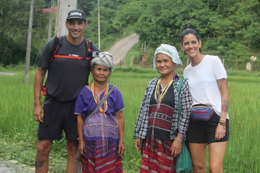 Four people standing in a rural area with green fields and a dirt road in the background. 1