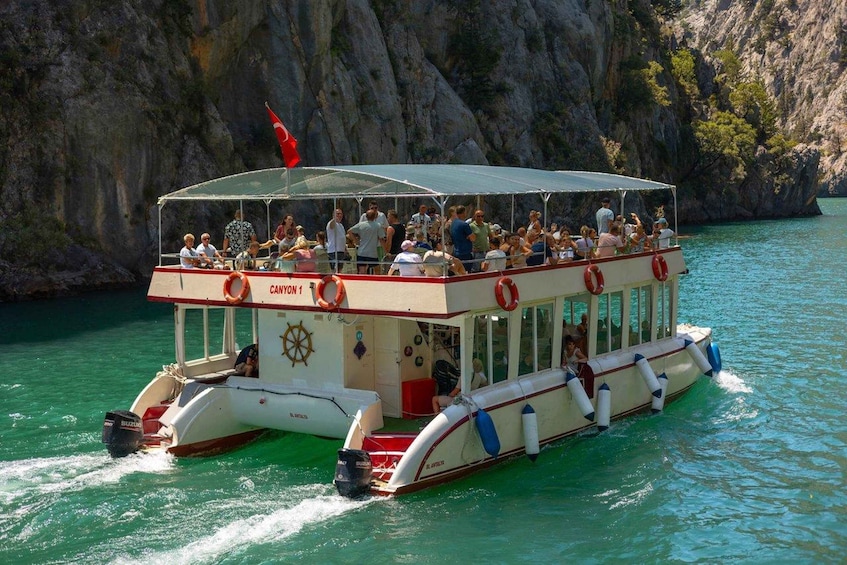 A crowded boat named 'CANYON 1' with a red flag on top, sailing on turquoise water with rocky cliffs in the background. 2