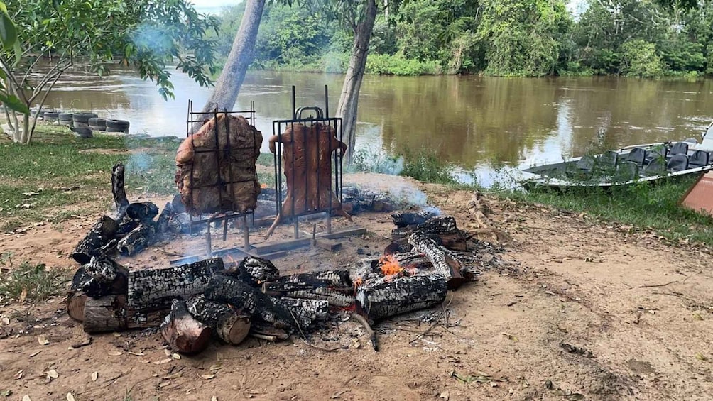 A campfire with a grill and a large pot over it, surrounded by logs, near a river. 3
