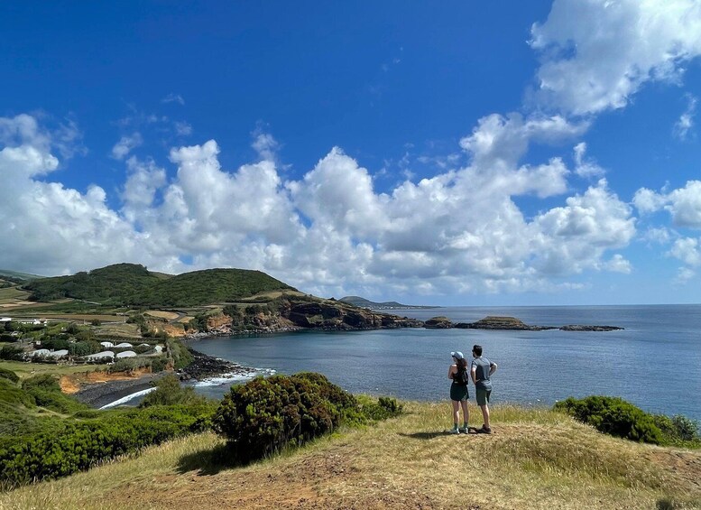Two people standing on a grassy hill overlooking a coastal landscape with hills, a body of water, and scattered houses. 1