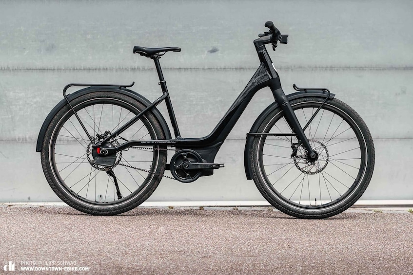 A black electric bicycle with a front basket and a rear rack, parked against a concrete wall. 4