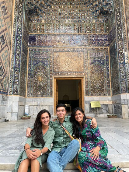 A group of people sitting in front of a building with intricate tile work and Arabic calligraphy. 1