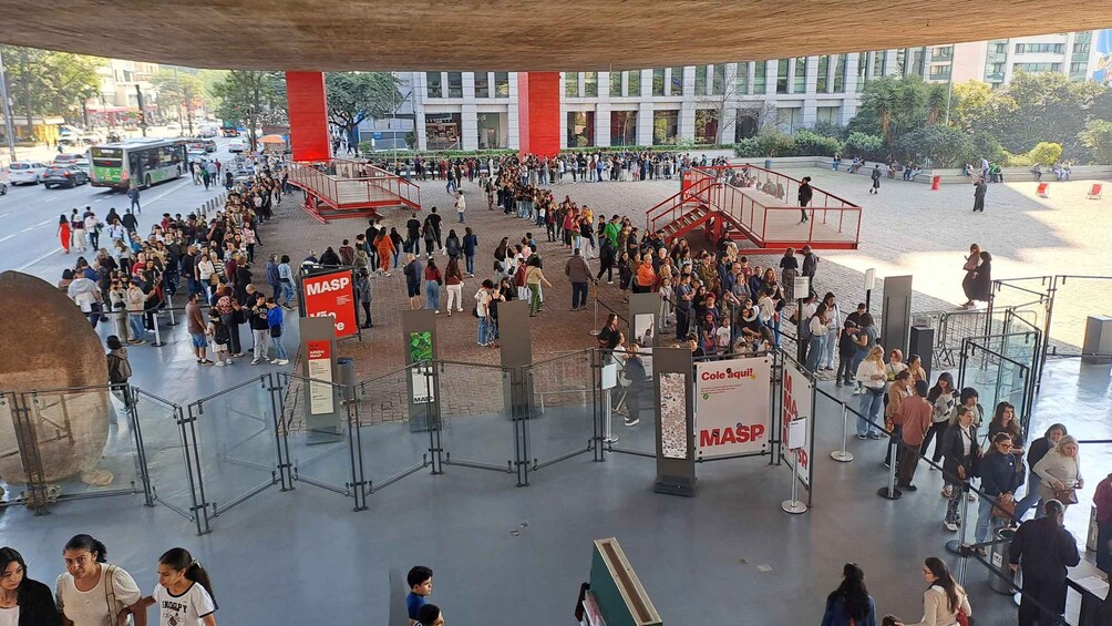 A crowded public square with people gathered around a central red structure, possibly a monument or information kiosk, with a modern building in the background. 4