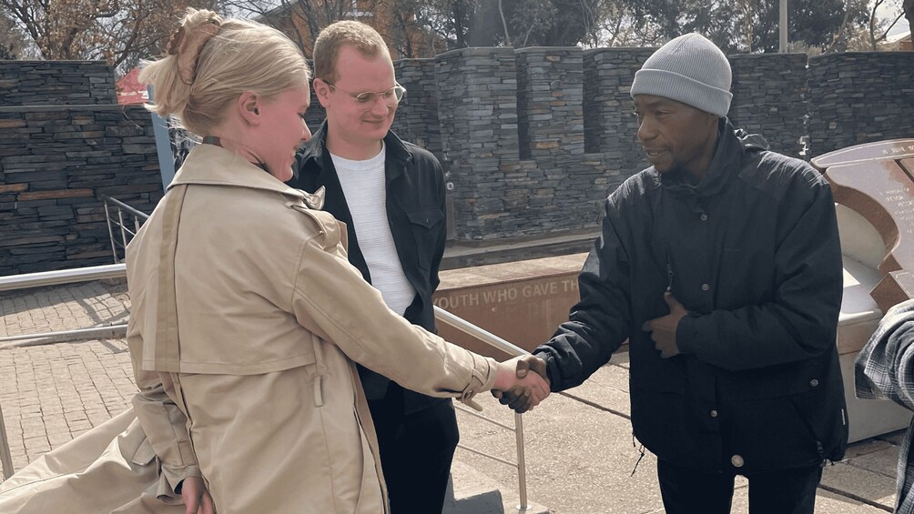 Three people shaking hands in front of a stone wall with a plaque. 5