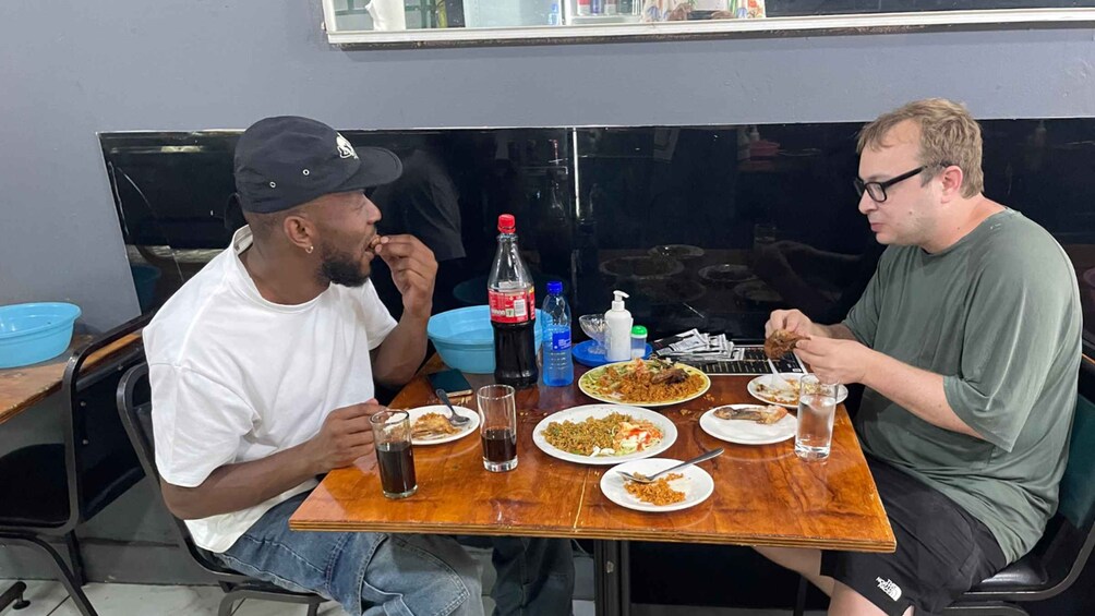 Two men sitting at a table with plates of food, drinks, and a bottle of soda. 1