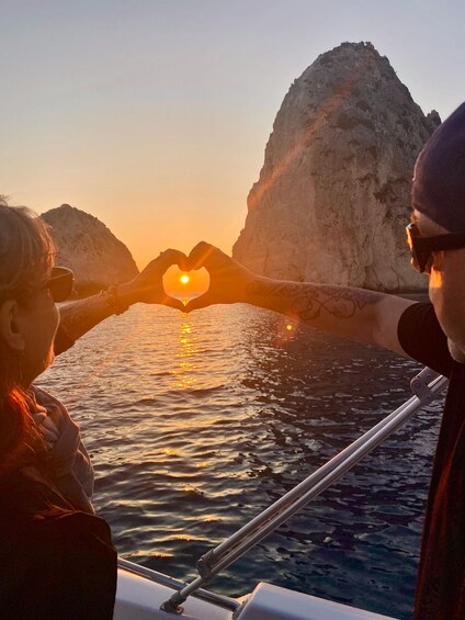 Two people making a heart shape with their hands, with the sun setting behind them on a body of water, surrounded by rocky cliffs. 3