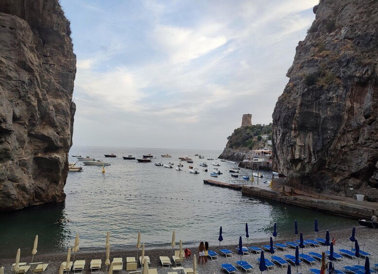 A rocky coastline with boats docked in the water, a tower on a cliff, and beach chairs and umbrellas along the shore. 4