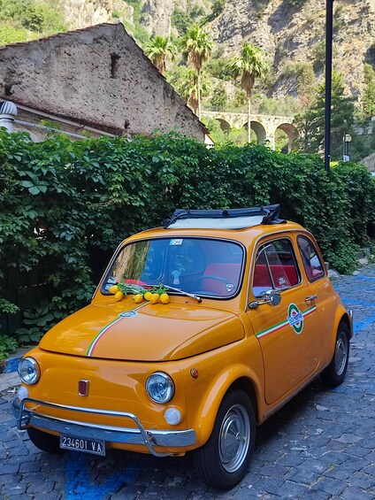 A vintage yellow car parked on a cobblestone street with lush greenery and a stone bridge in the background. 2