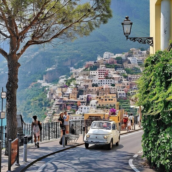 A coastal town with buildings on a hillside, a vintage car on the road, and pedestrians walking along the sidewalk. 3