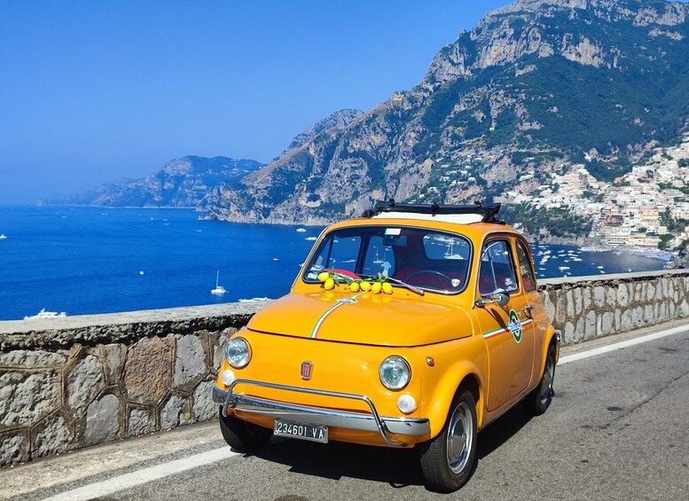 A yellow vintage car parked on a coastal road with a scenic view of the sea and mountains in the background. 1