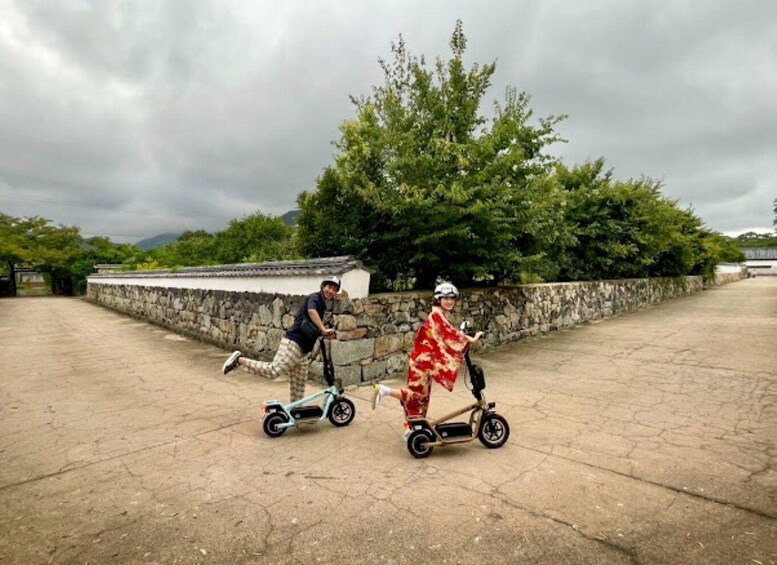 Two people riding electric scooters on a paved area with stone walls and trees in the background. 4