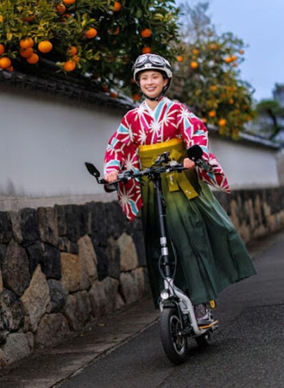 A person riding a scooter on a paved path with a stone wall and orange trees in the background. 1