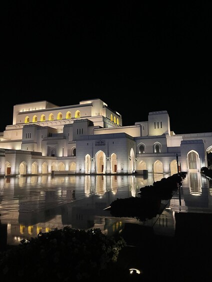 A large, illuminated building with arched windows and a reflective water surface in front. 1