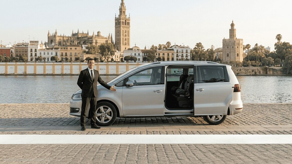 A man in a suit standing next to a silver minivan with its door open, parked by a river with historic buildings in the background. 3