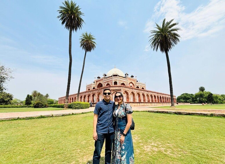 A couple standing in front of a large, historic building with a dome and arched windows, surrounded by palm trees and a well-maintained lawn. 1