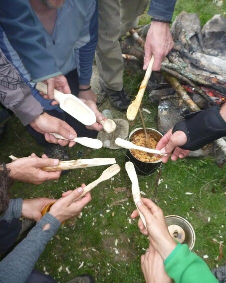People holding wooden utensils around a pot of food, outdoors near a campfire. 4