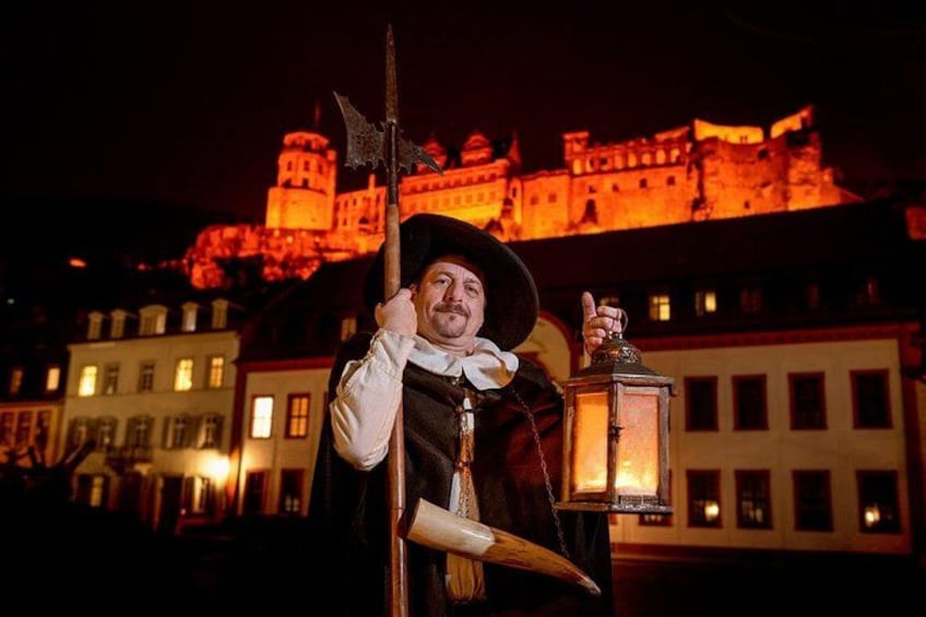 Eine Person in historischer Tracht hält eine Laterne und einen Stab, im Hintergrund ist ein Schloss zu sehen. 3