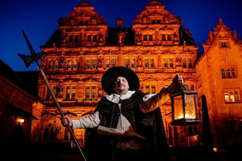 Eine Person in historischer Tracht mit einer Laterne steht vor einem großen, reich verzierten Gebäude bei Nacht. 1