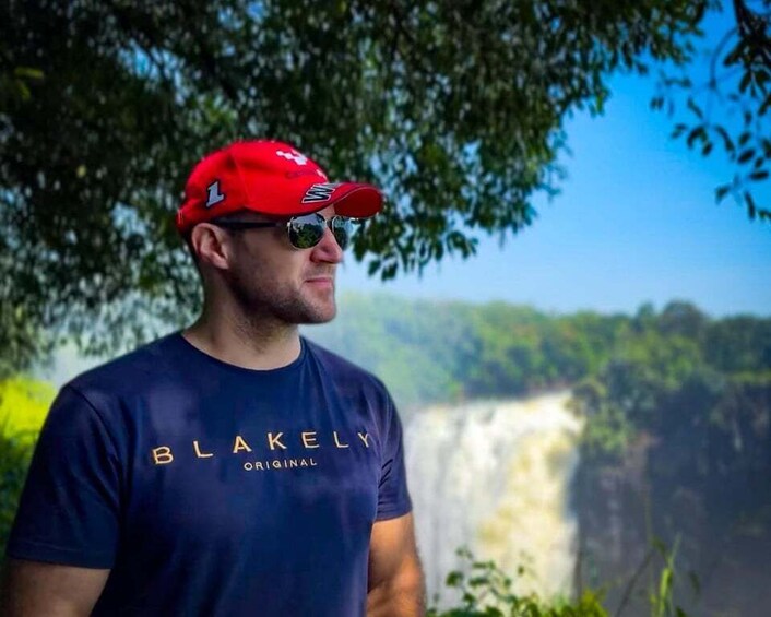 A person wearing a red cap and a blue t-shirt with 'BLAKELY ORIGINAL' text, standing in front of a waterfall. 2