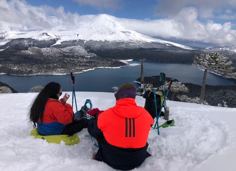 Two people sitting on snow-covered ground, facing a snow-capped mountain and a lake. 5