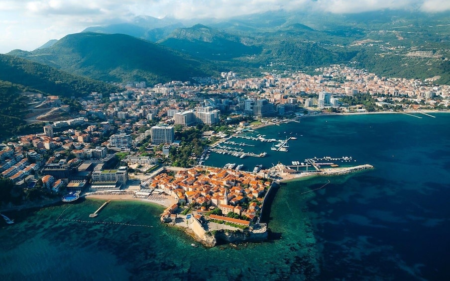 An aerial view of a coastal city with a historic walled town, a marina, and surrounding mountains. 4