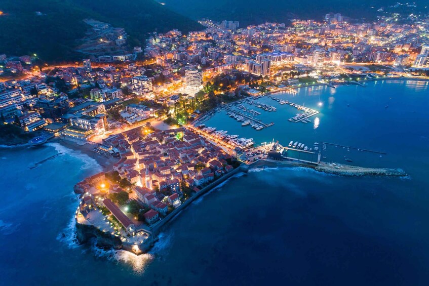 An aerial view of a coastal city at night with illuminated buildings, a marina, and a fortified area along the shore. 3