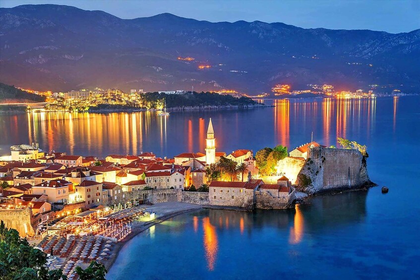 A coastal town with a prominent tower, a fortified structure, and a calm sea reflecting city lights. 1