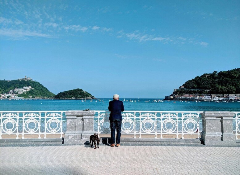 A man and a dog on a paved promenade overlooking a coastal town with boats and hills. 1