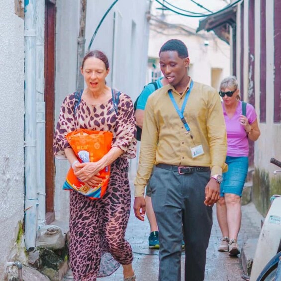A narrow street with buildings on either side, a person carrying a bag, and another person walking beside them. 4