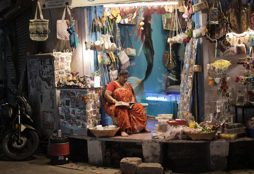 A woman sitting at a stall with various items for sale, including bags and jewelry. 4