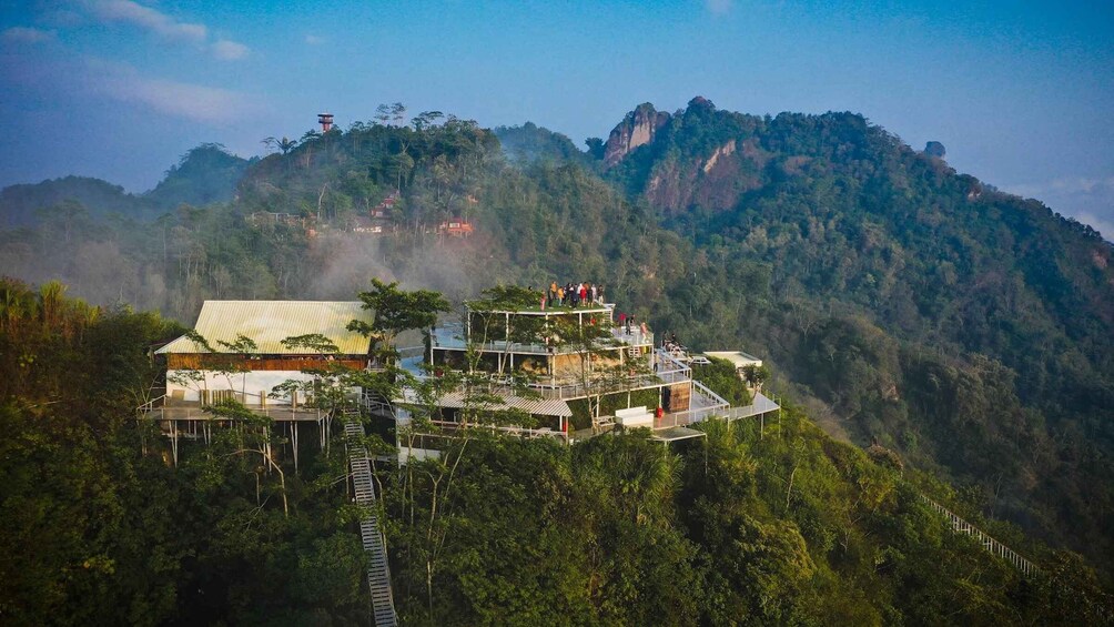 A multi-level wooden structure with a metal roof, surrounded by dense forest and a mountain in the background. 3