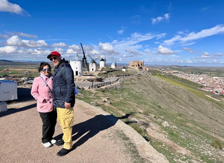 A couple standing on a hill overlooking windmills and a historic castle. 1