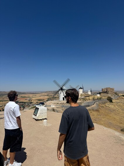 Two people standing on a hill overlooking windmills and a castle. 3