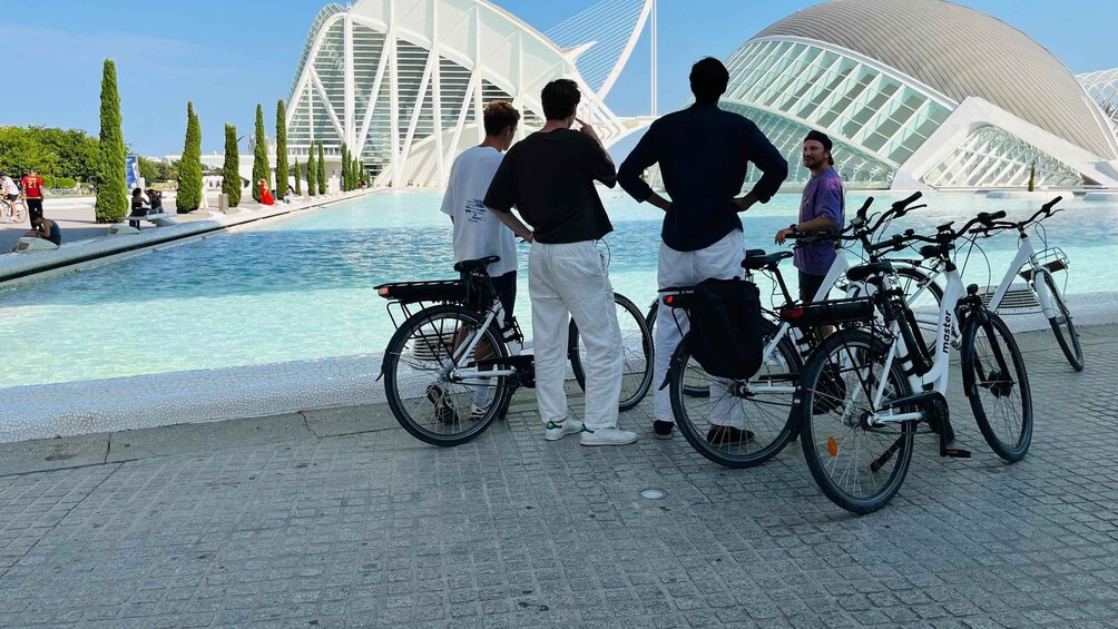 People standing by a waterfront with bicycles, a modern architectural structure in the background, and a clear sky. 4