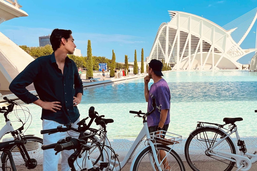Two people conversing near bicycles by a waterfront with a distinctive white bridge in the background. 1