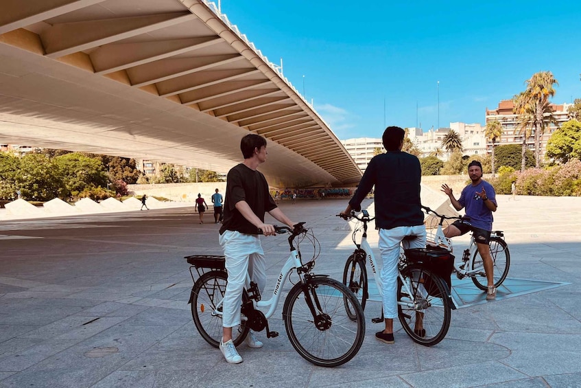 Three people on bicycles under a bridge, with a cityscape in the background. 2