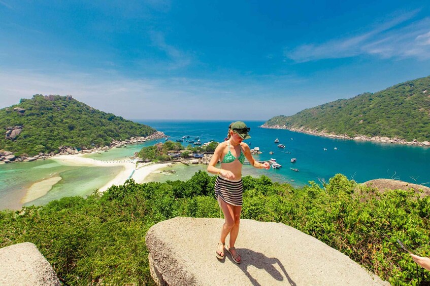 A woman in a bikini standing on a rock overlooking a coastal landscape with boats and a beach. 1