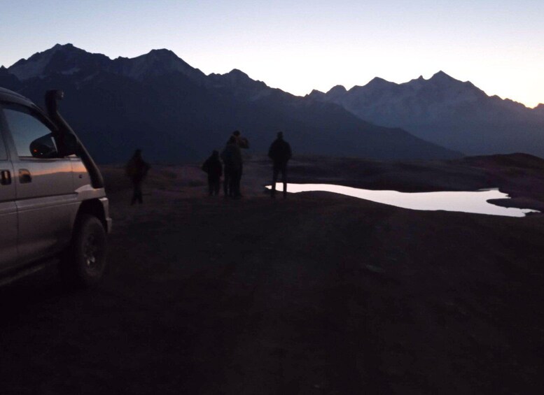 A group of people standing near a vehicle and a small body of water in a mountainous area. 5